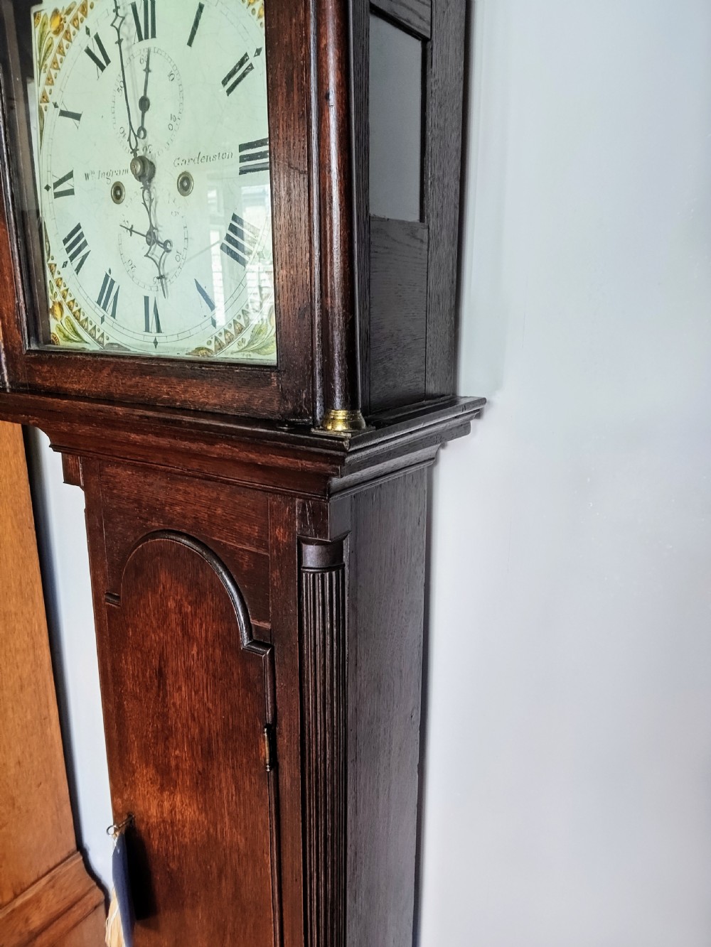 An antique Georgian dark oak grandfather clock featuring a swan-neck pediment, fluted columns, an arched trunk door, and a hand-painted floral dial. View VI