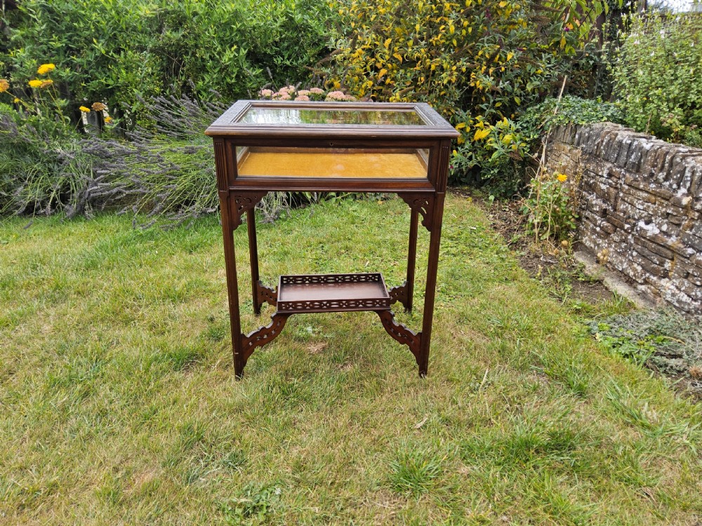 A rectangular dark mahogany bijouterie table featuring glazed sides and top, a gold fabric lining inside, pierced fretwork corner spandrels, and a lower tier shelf with a fretwork gallery.