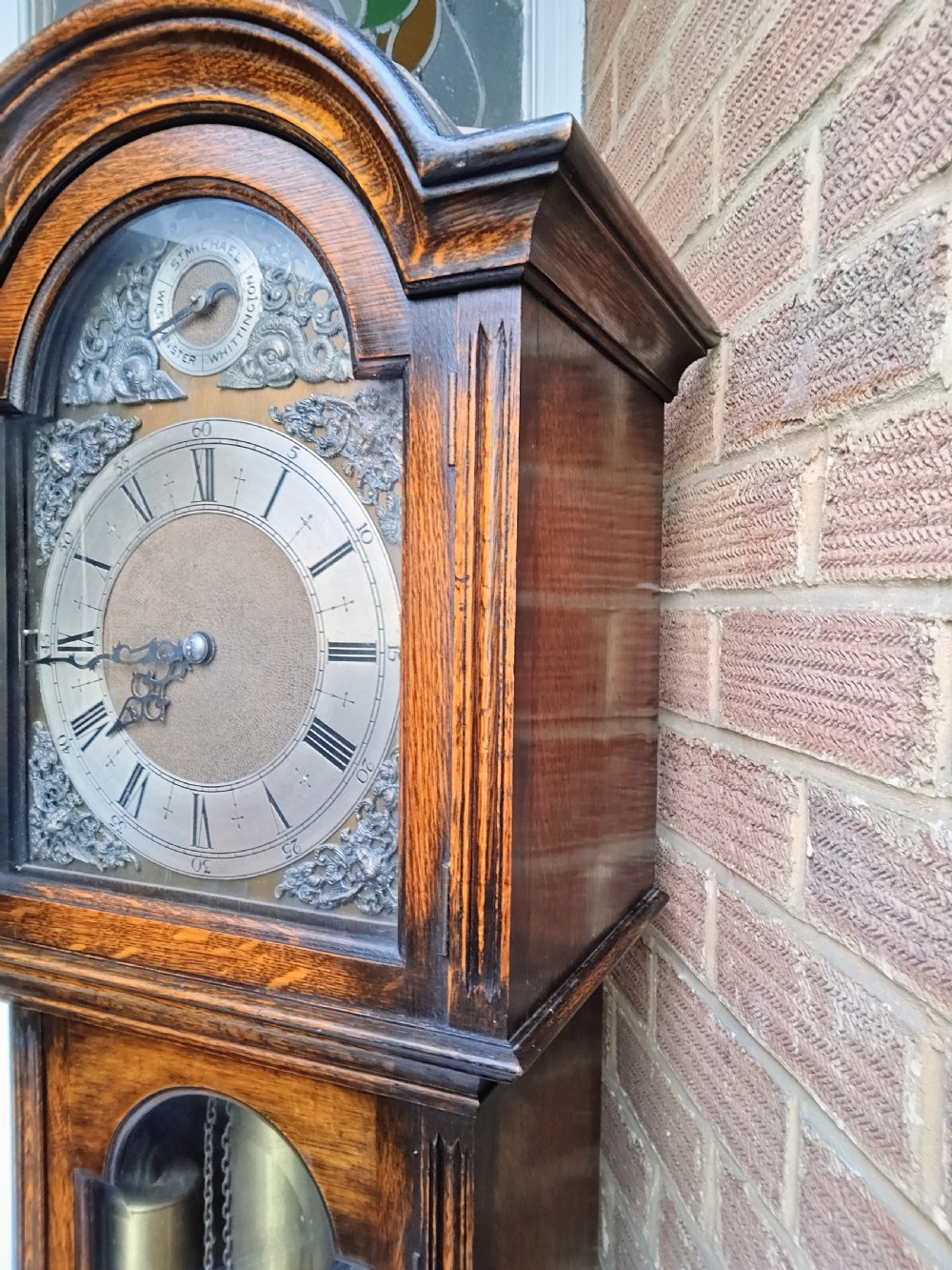 A traditional warm oak grandmother longcase clock featuring an arched top hood, silvered dial, glazed door revealing brass weights and pendulum, and a solid base. View IV