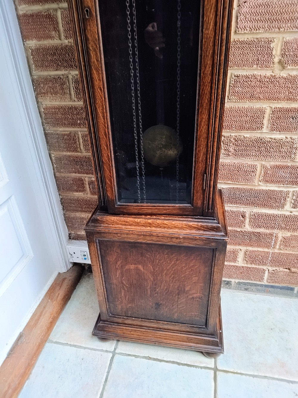 A traditional warm oak grandmother longcase clock featuring an arched top hood, silvered dial, glazed door revealing brass weights and pendulum, and a solid base. View VIII