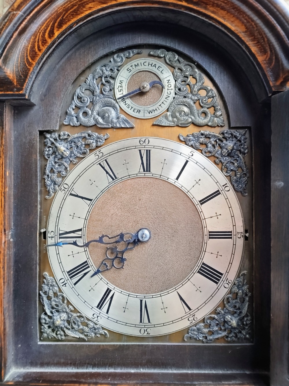 A traditional warm oak grandmother longcase clock featuring an arched top hood, silvered dial, glazed door revealing brass weights and pendulum, and a solid base. View X