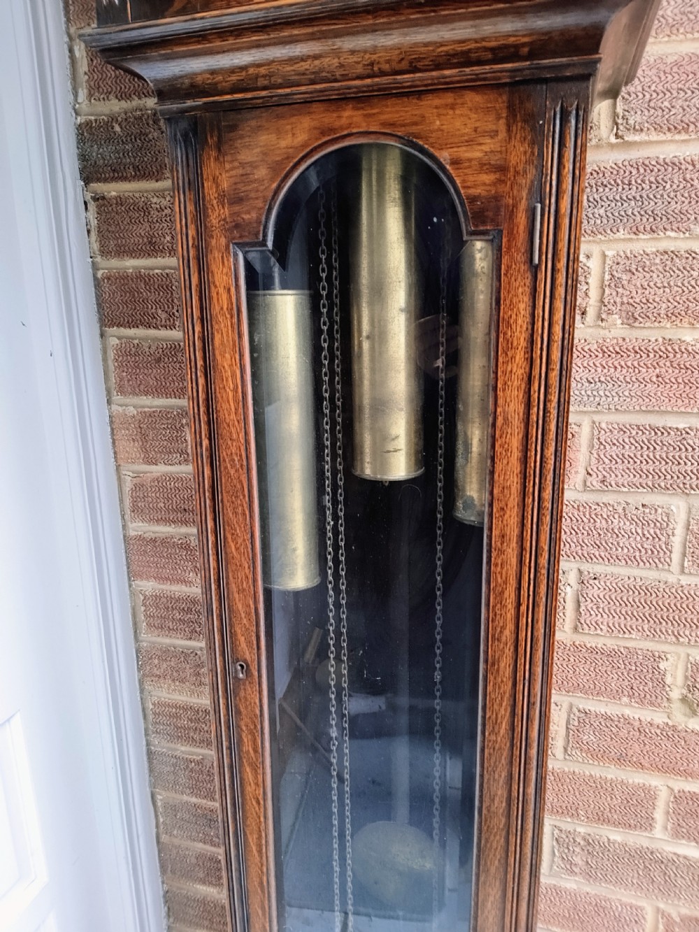 A traditional warm oak grandmother longcase clock featuring an arched top hood, silvered dial, glazed door revealing brass weights and pendulum, and a solid base. View XI