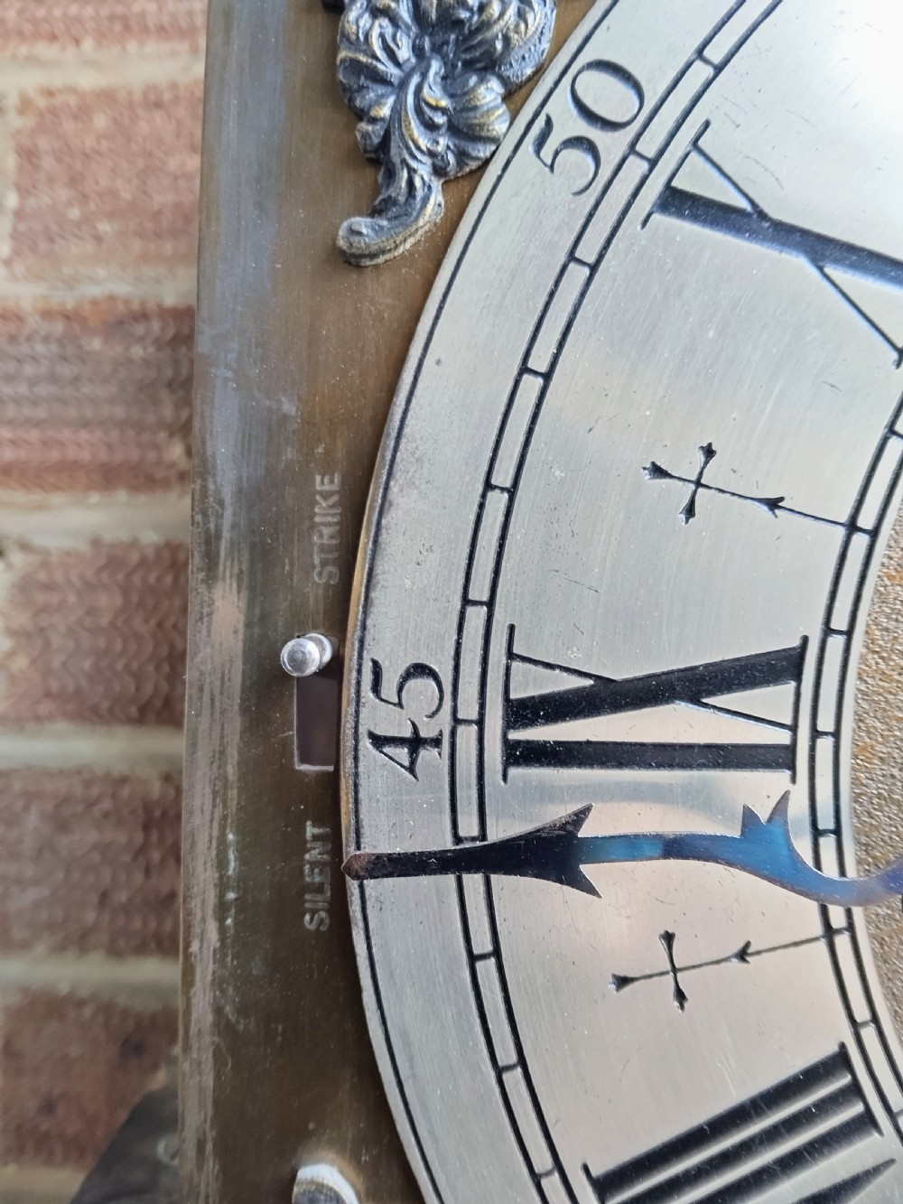 A traditional warm oak grandmother longcase clock featuring an arched top hood, silvered dial, glazed door revealing brass weights and pendulum, and a solid base. View XII