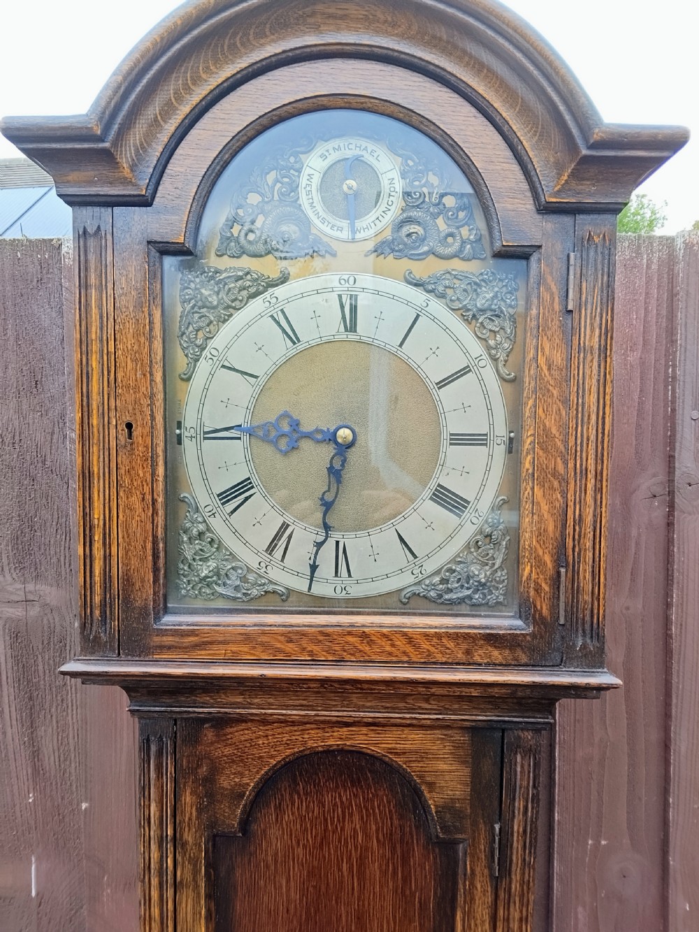 An oak grandmother longcase clock featuring an arched top, a brass dial with Roman numerals, and a tall wooden casing. View II