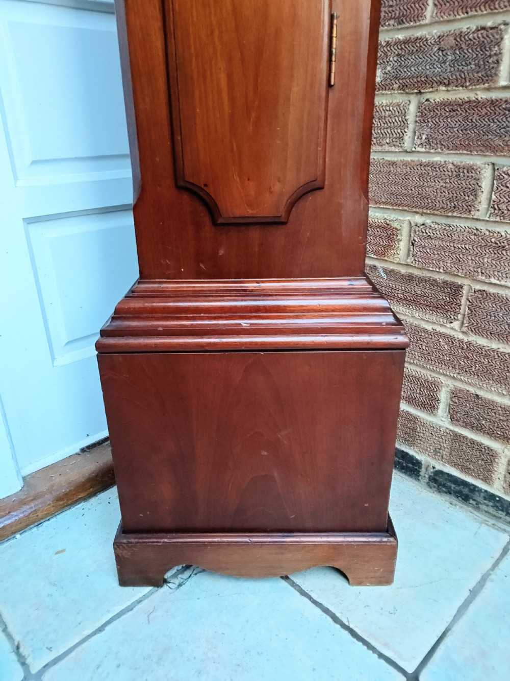 A slim mahogany grandmother longcase clock with a carved top frieze, brass Roman numeral dial, and an arched waist door. View II