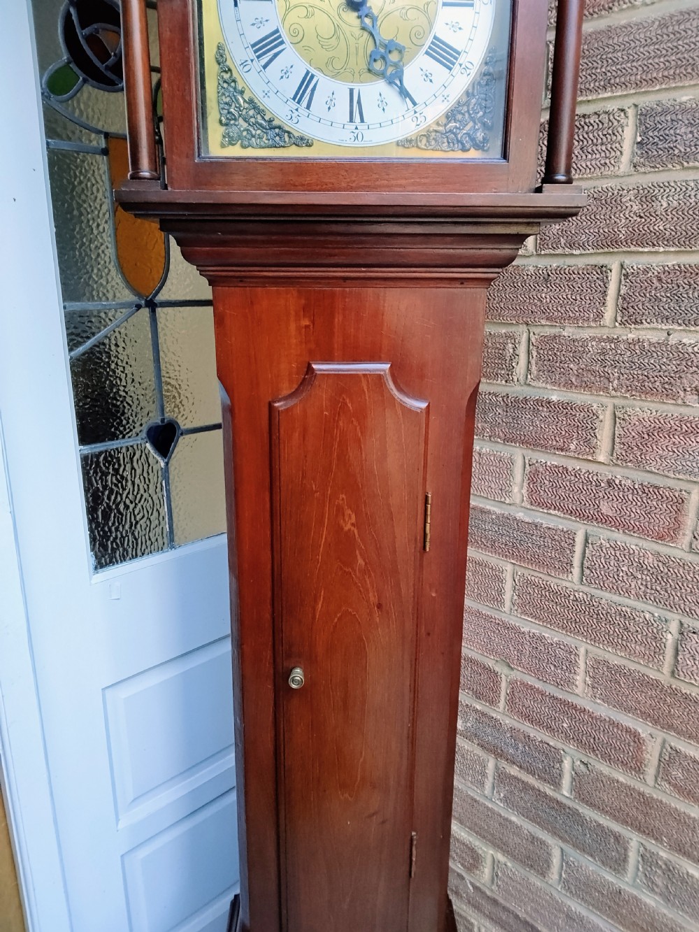 A slim mahogany grandmother longcase clock with a carved top frieze, brass Roman numeral dial, and an arched waist door. View IV