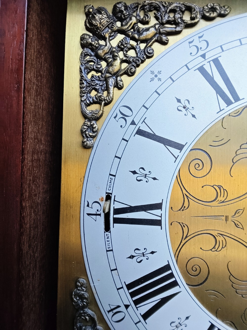 A slim mahogany grandmother longcase clock with a carved top frieze, brass Roman numeral dial, and an arched waist door. View VI