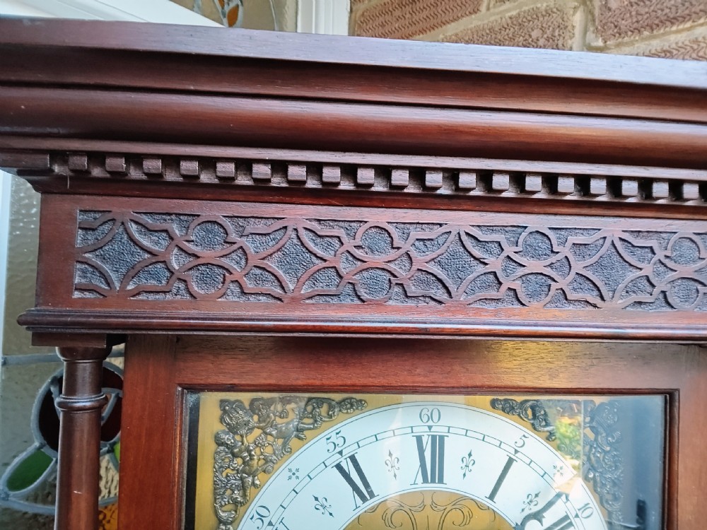 A slim mahogany grandmother longcase clock with a carved top frieze, brass Roman numeral dial, and an arched waist door. View VIII