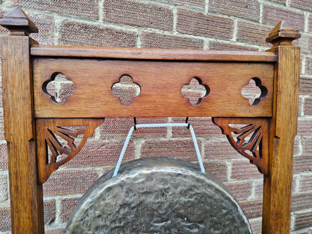 A freestanding Gothic Revival oak frame containing carved trefoil cut-outs, suspending a central circular hammered brass dinner gong with a wooden striker. View III