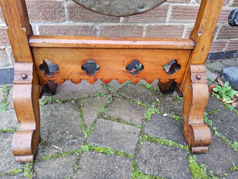 A freestanding Gothic Revival oak frame containing carved trefoil cut-outs, suspending a central circular hammered brass dinner gong with a wooden striker. View IV