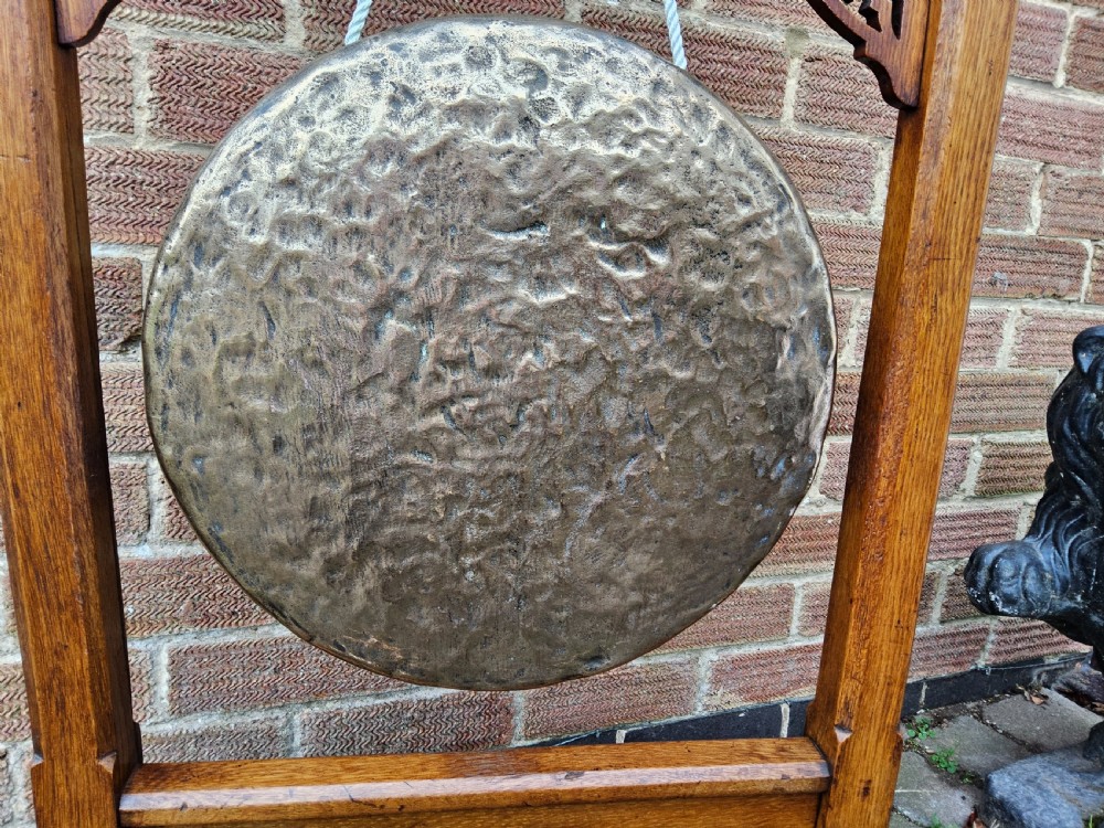 A freestanding Gothic Revival oak frame containing carved trefoil cut-outs, suspending a central circular hammered brass dinner gong with a wooden striker. View V