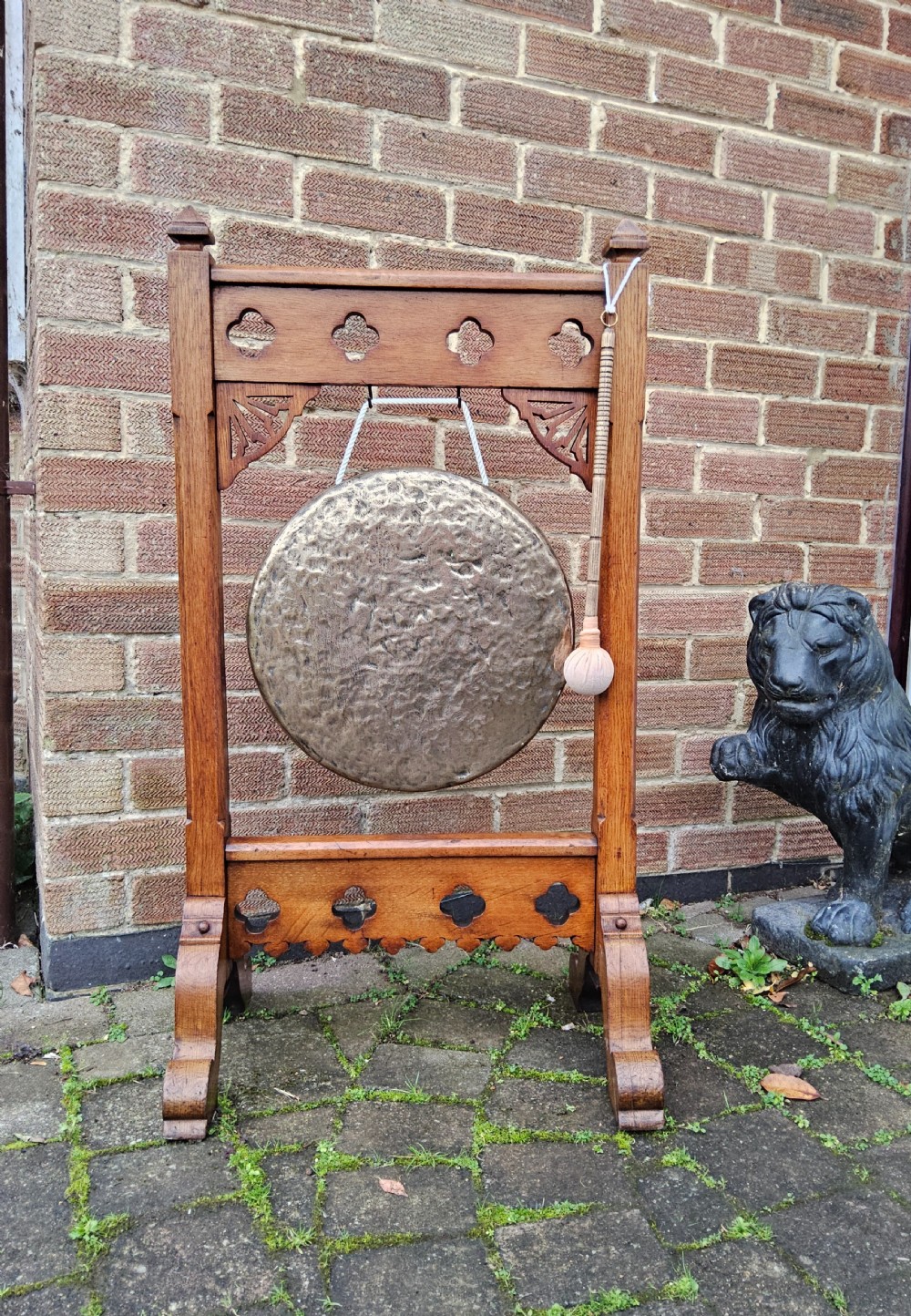 A freestanding Gothic Revival oak frame containing carved trefoil cut-outs, suspending a central circular hammered brass dinner gong with a wooden striker. View VII