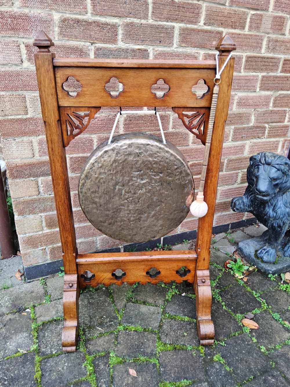 A freestanding Gothic Revival oak frame containing carved trefoil cut-outs, suspending a central circular hammered brass dinner gong with a wooden striker.