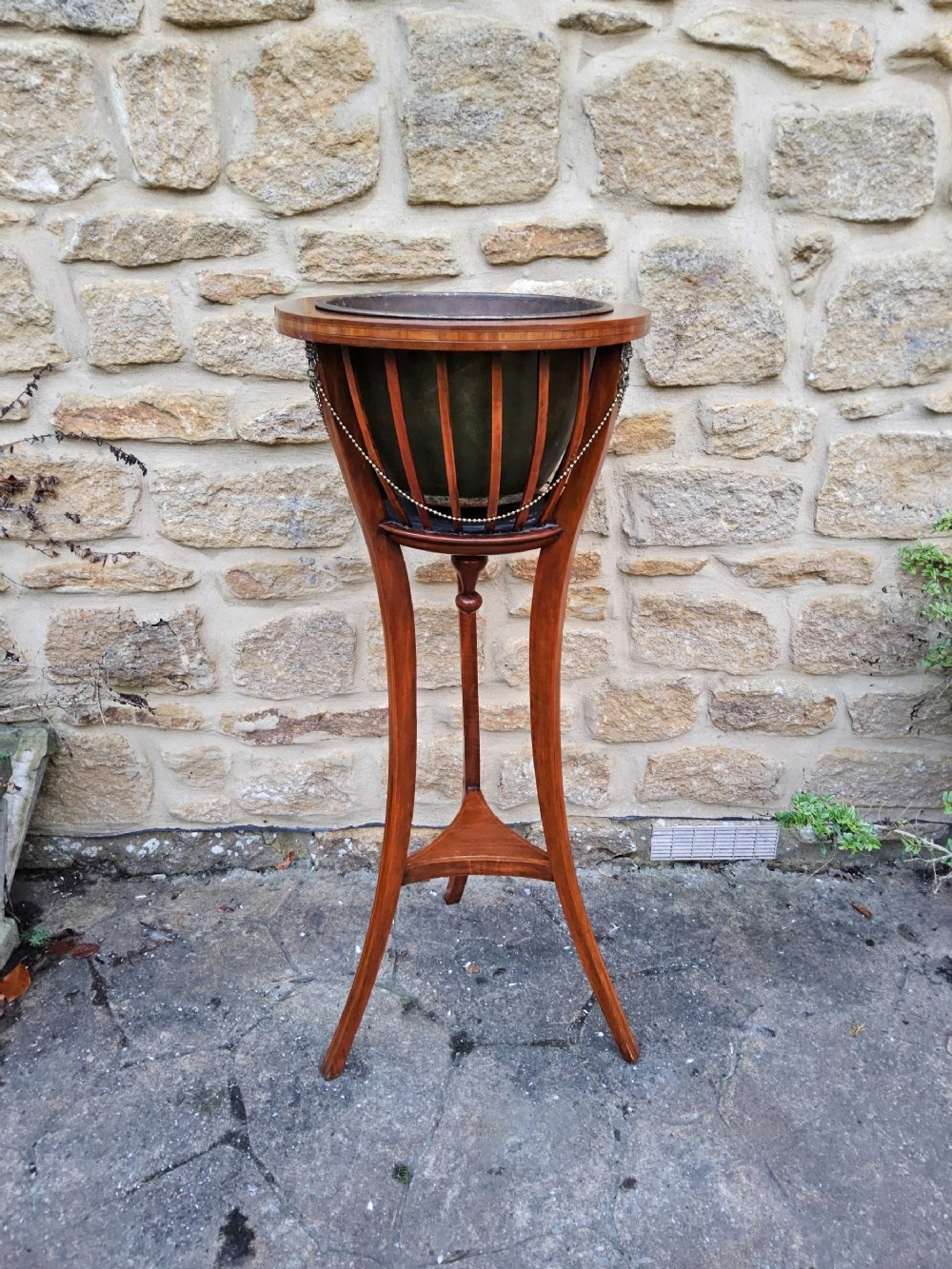 An Edwardian mahogany tripod jardiniere stand featuring slatted sides, satinwood stringing inlay around the top rim, three sweeping splayed legs, and a removable metal liner.