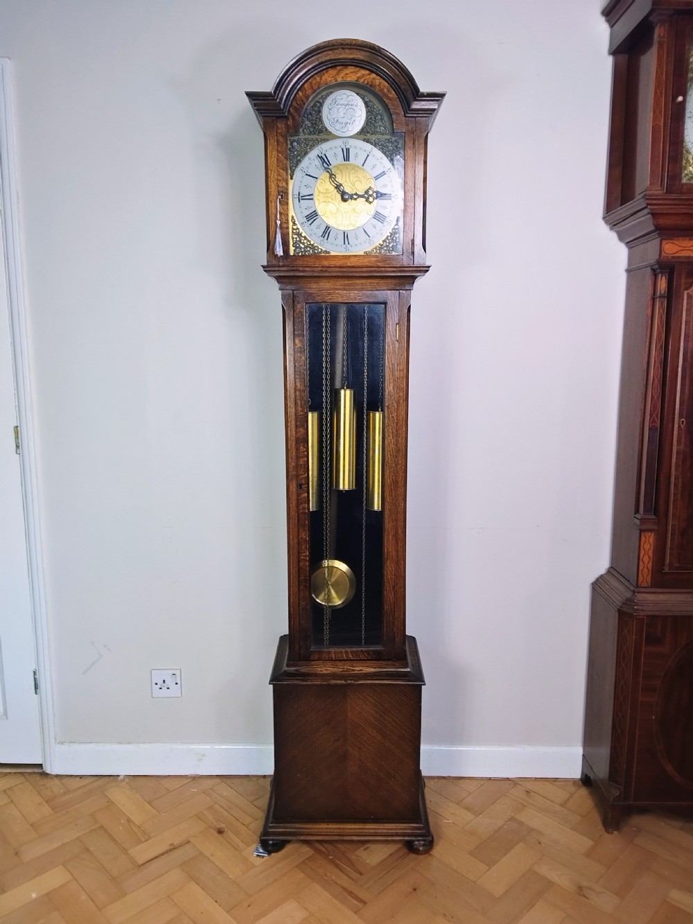 An English oak longcase clock featuring an arched hood, silvered brass dial, and a glazed trunk door displaying three brass weights and a pendulum. View IX