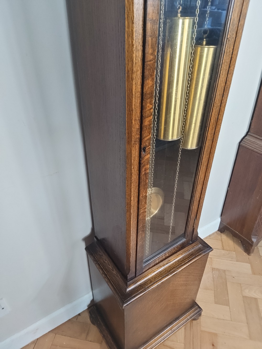 An English oak longcase clock featuring an arched hood, silvered brass dial, and a glazed trunk door displaying three brass weights and a pendulum. View IV