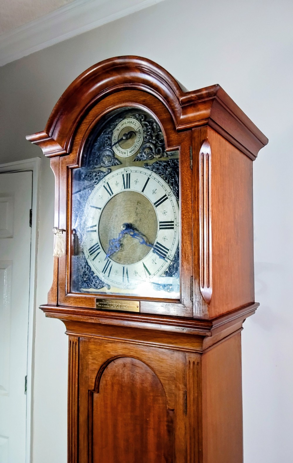 A tall mahogany longcase clock featuring an arched hood, glass door displaying a brass dial with silvered chapter ring, fluted side columns, and a moulded plinth base. View IV