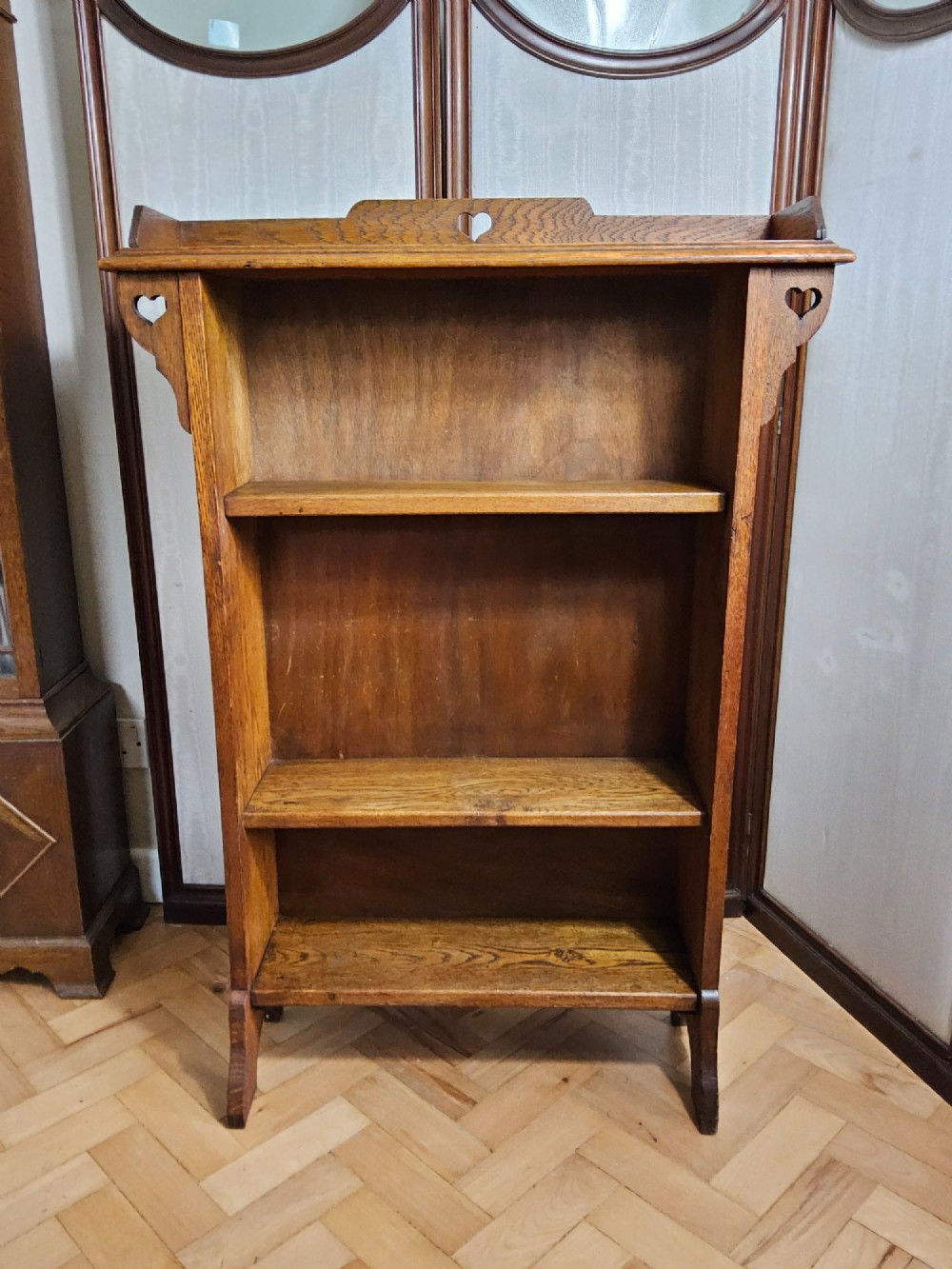 An Arts and Crafts solid oak open bookcase featuring three shelves and heart-shaped cutouts on the top gallery and side brackets.