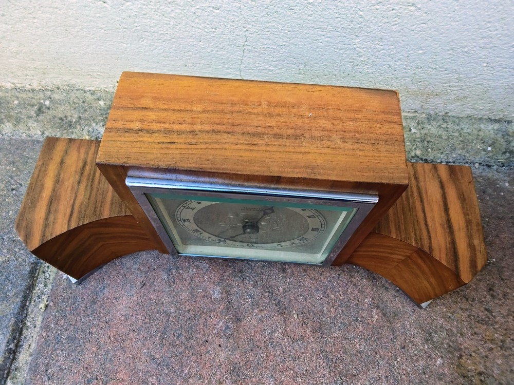An Art Deco walnut mantel clock featuring chevron-patterned wooden wings, a central square face with Roman numerals, and chrome trim accents. View II