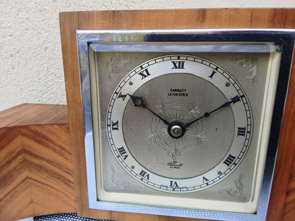 An Art Deco walnut mantel clock featuring chevron-patterned wooden wings, a central square face with Roman numerals, and chrome trim accents. View VI