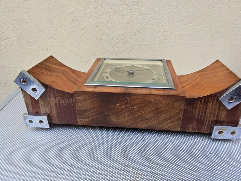 An Art Deco walnut mantel clock featuring chevron-patterned wooden wings, a central square face with Roman numerals, and chrome trim accents. View IX
