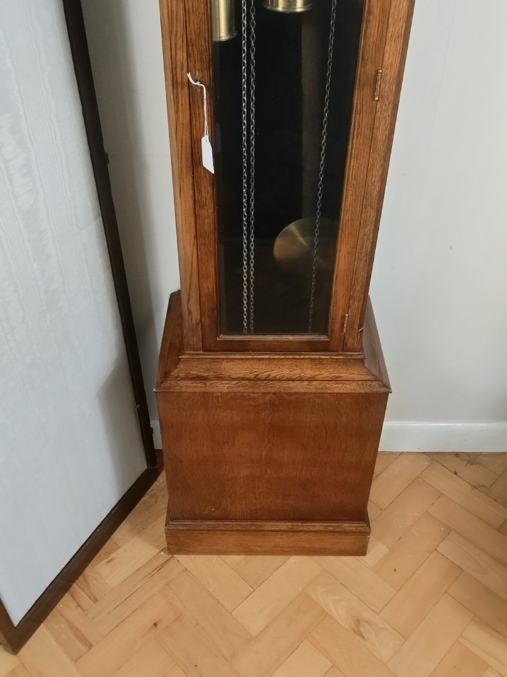 A freestanding wooden longcase clock featuring an arched hood, a brass and silvered dial, and a glazed trunk door revealing weights and a pendulum. View V