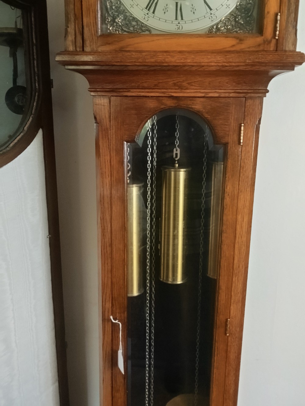 A freestanding wooden longcase clock featuring an arched hood, a brass and silvered dial, and a glazed trunk door revealing weights and a pendulum. View VIII