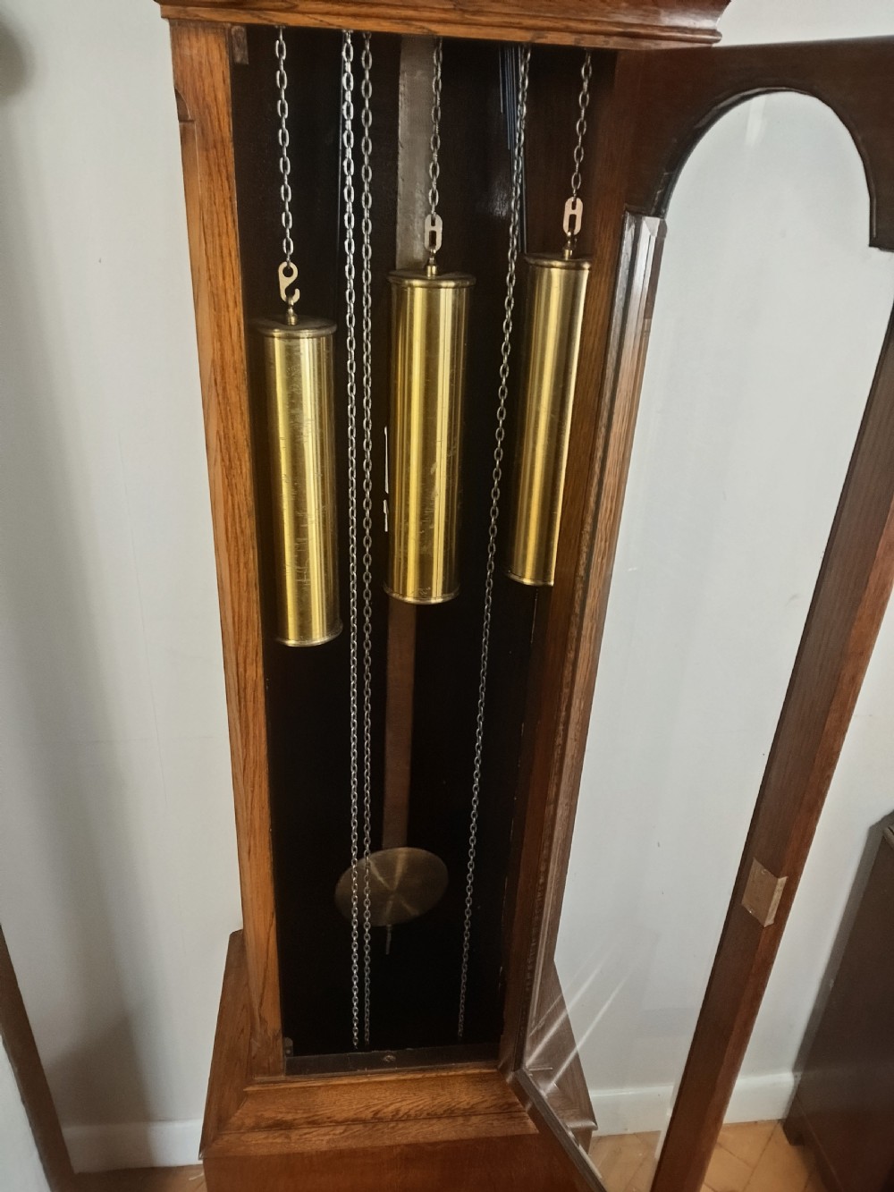 A freestanding wooden longcase clock featuring an arched hood, a brass and silvered dial, and a glazed trunk door revealing weights and a pendulum. View X