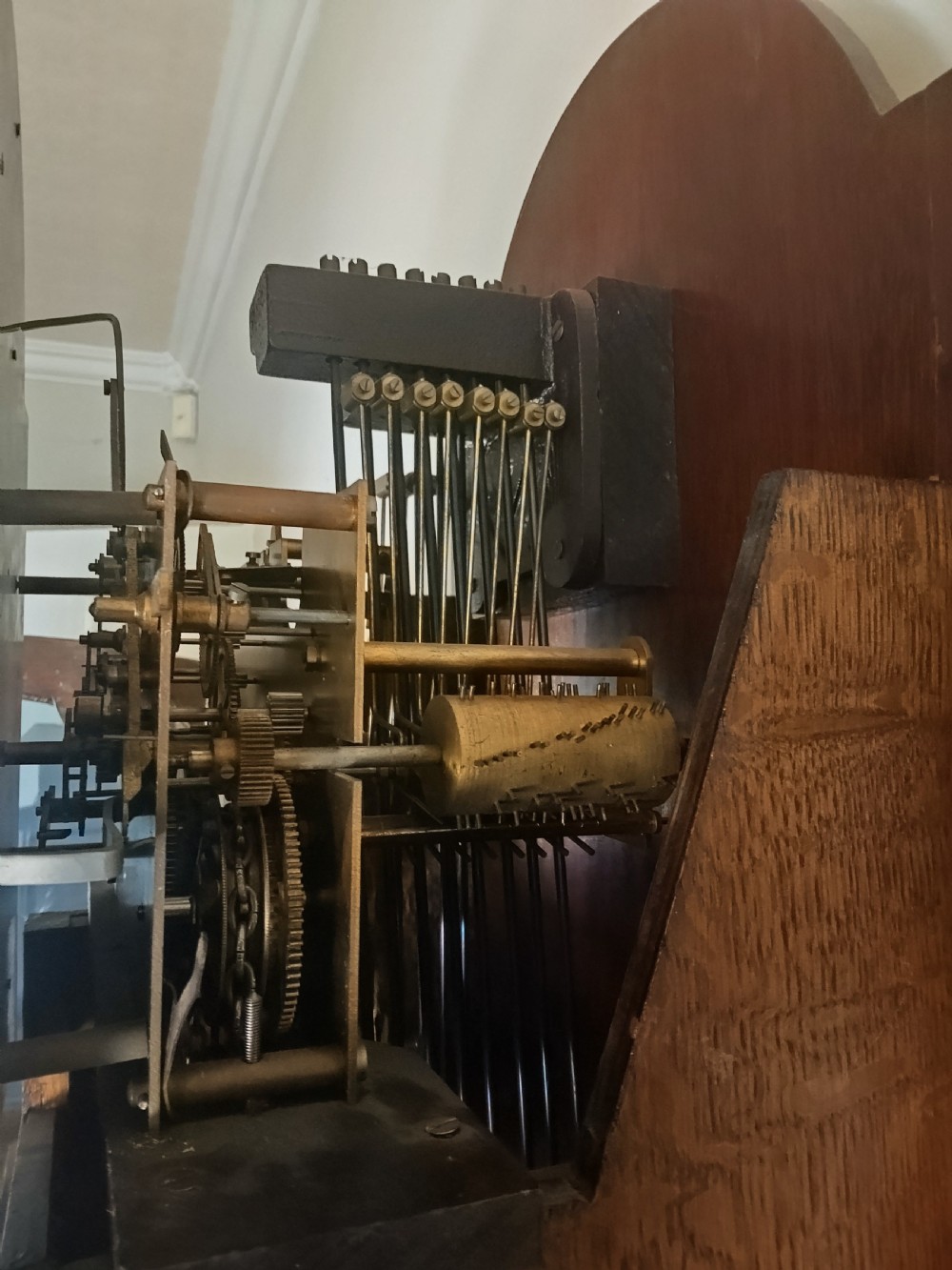 A freestanding wooden longcase clock featuring an arched hood, a brass and silvered dial, and a glazed trunk door revealing weights and a pendulum. View XII