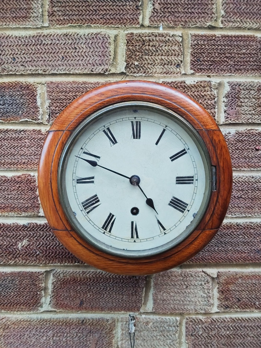 A circular late nineteenth-century oak dial wall clock featuring an off-white face with Roman numerals and a brass bezel.