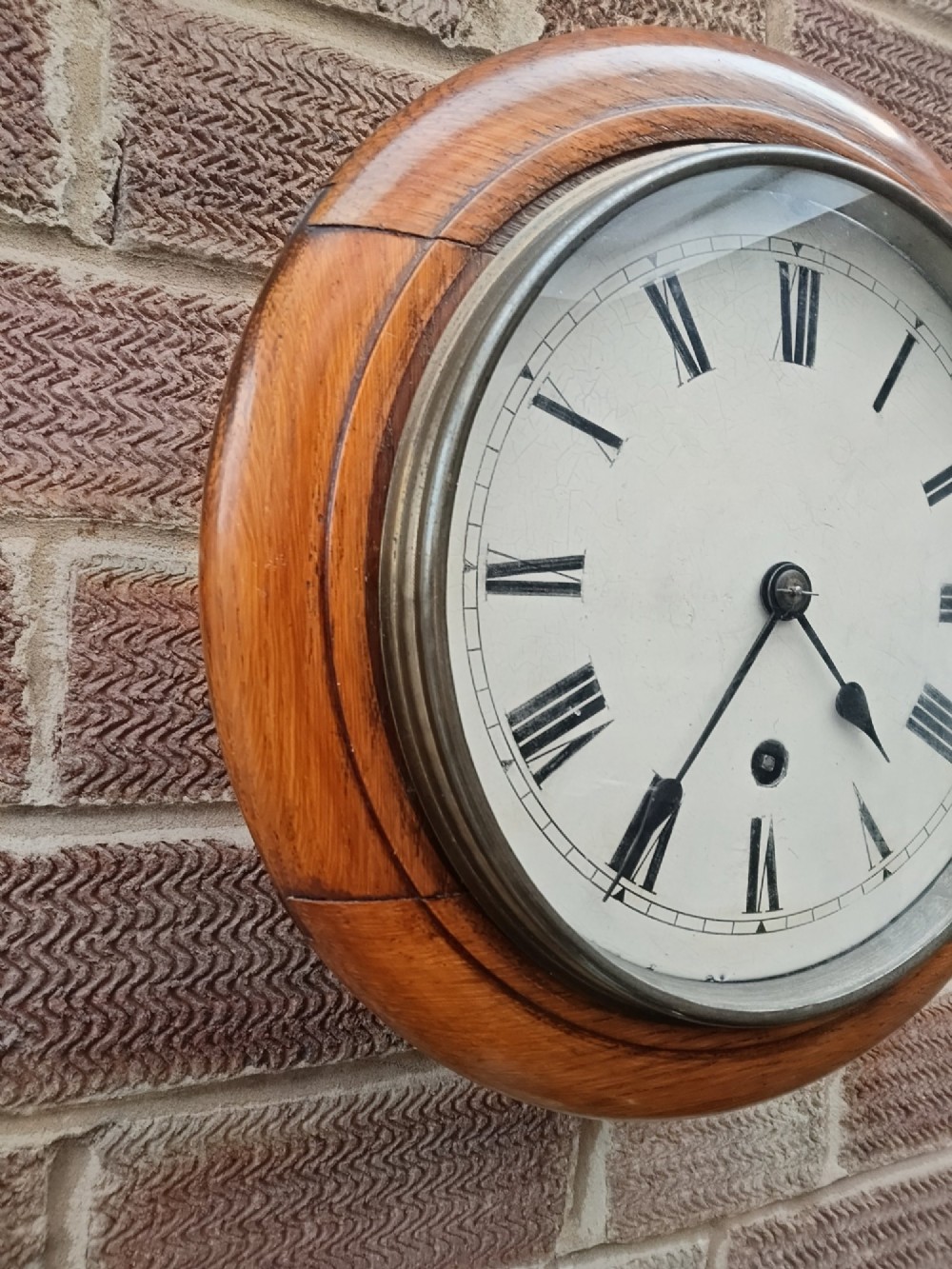A circular late nineteenth-century oak dial wall clock featuring an off-white face with Roman numerals and a brass bezel. View IV