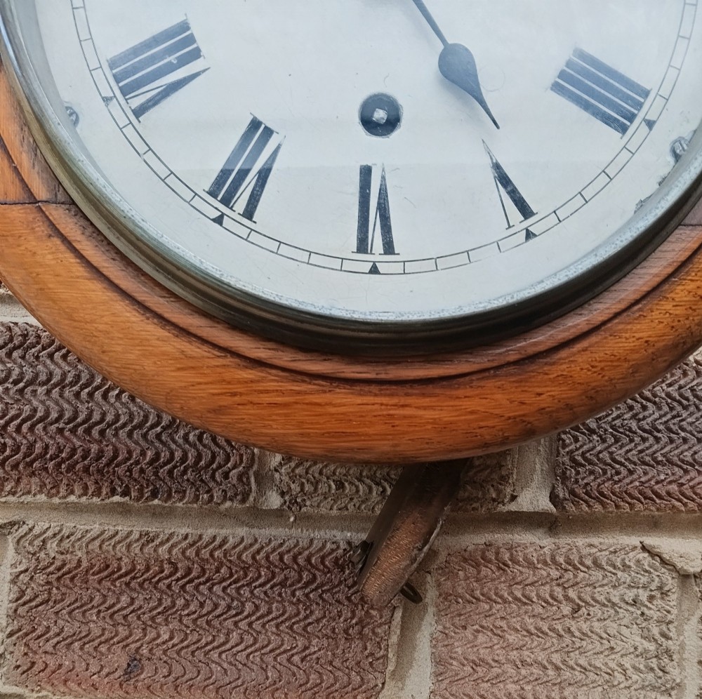 A circular late nineteenth-century oak dial wall clock featuring an off-white face with Roman numerals and a brass bezel. View VI