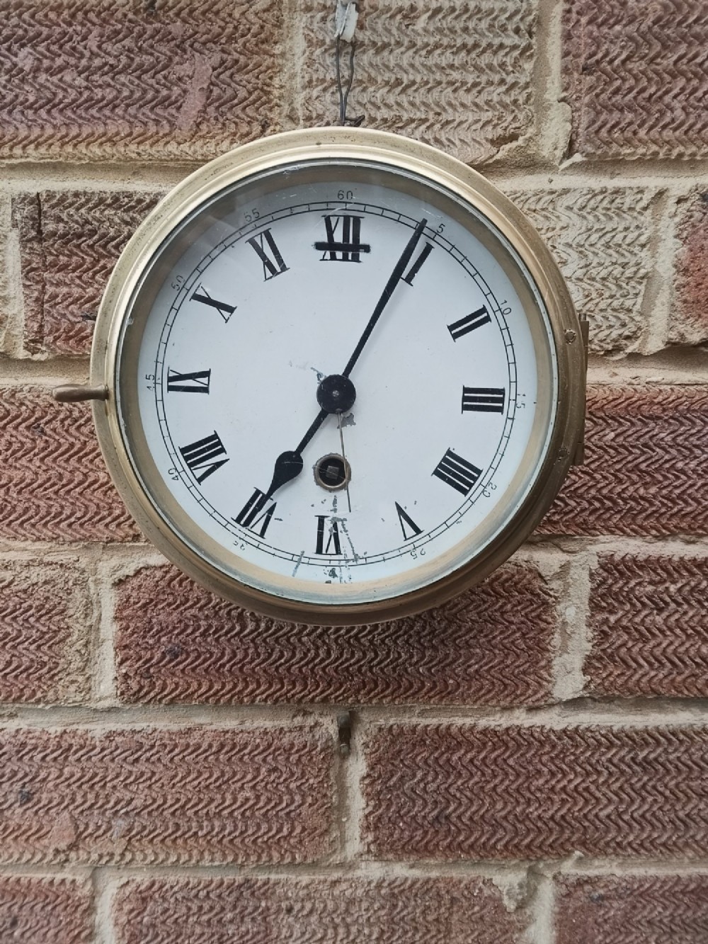 A circular antique brass marine bulkhead wall clock featuring a white dial with black Roman numerals and side latch fixtures.