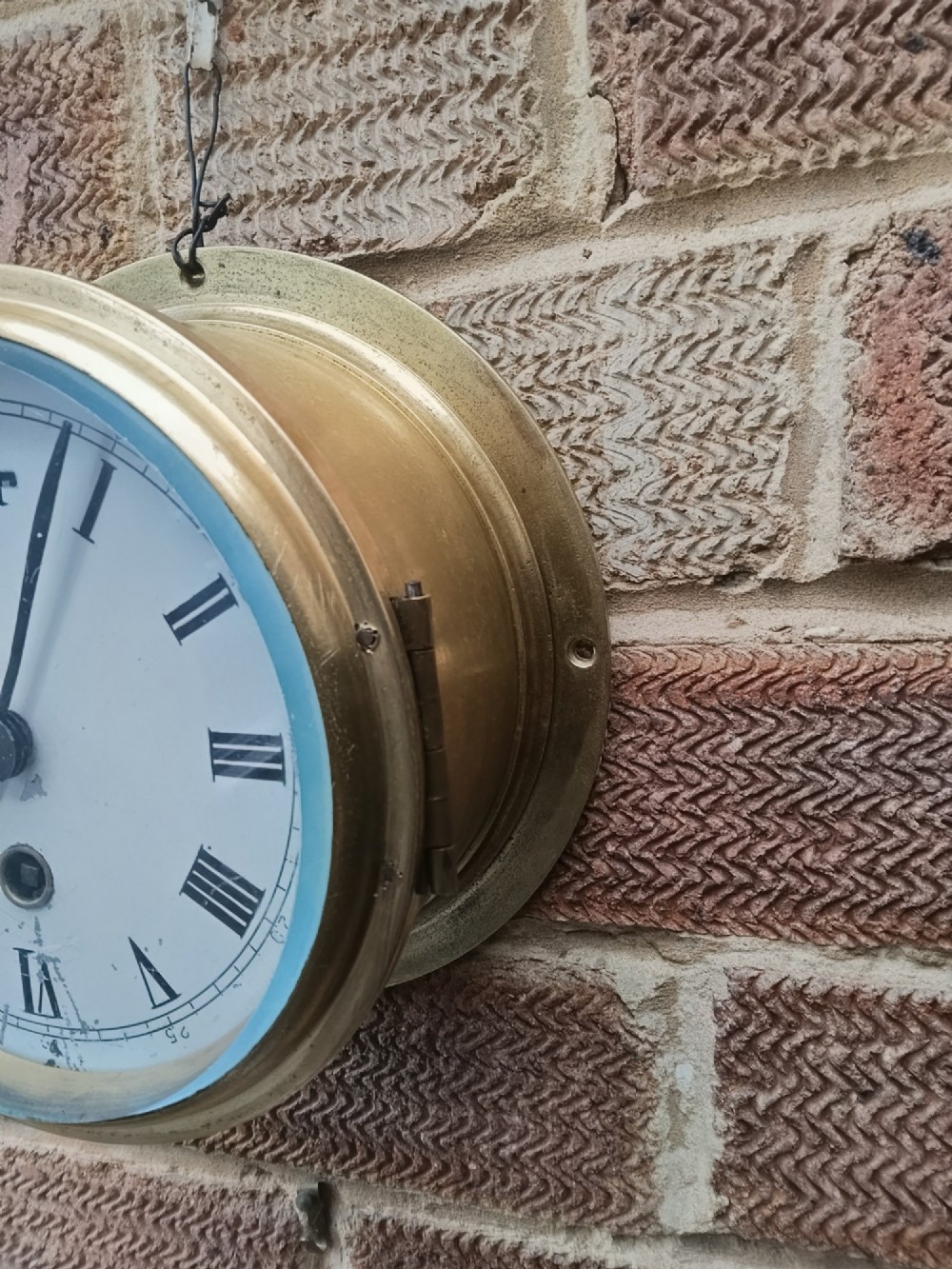 A circular antique brass marine bulkhead wall clock featuring a white dial with black Roman numerals and side latch fixtures. View II