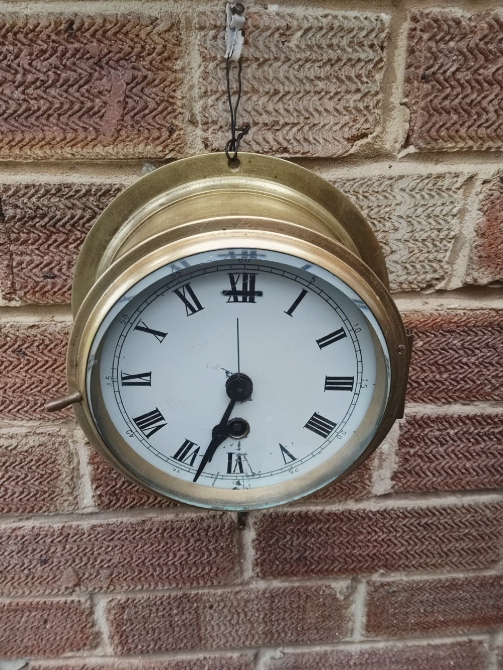 A circular antique brass marine bulkhead wall clock featuring a white dial with black Roman numerals and side latch fixtures. View IV