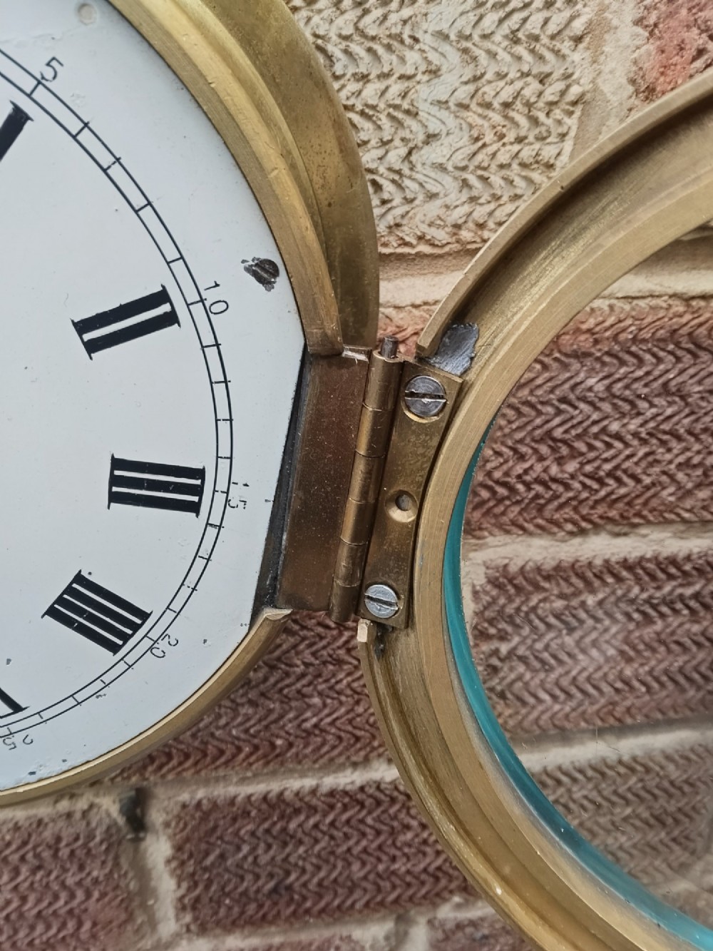 A circular antique brass marine bulkhead wall clock featuring a white dial with black Roman numerals and side latch fixtures. View V