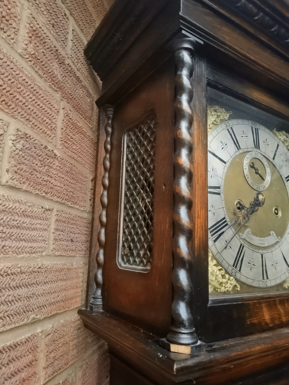 A tall dark oak longcase clock featuring rope twist columns, a square brass dial with silvered chapter ring, and a carved wooden casing. View V
