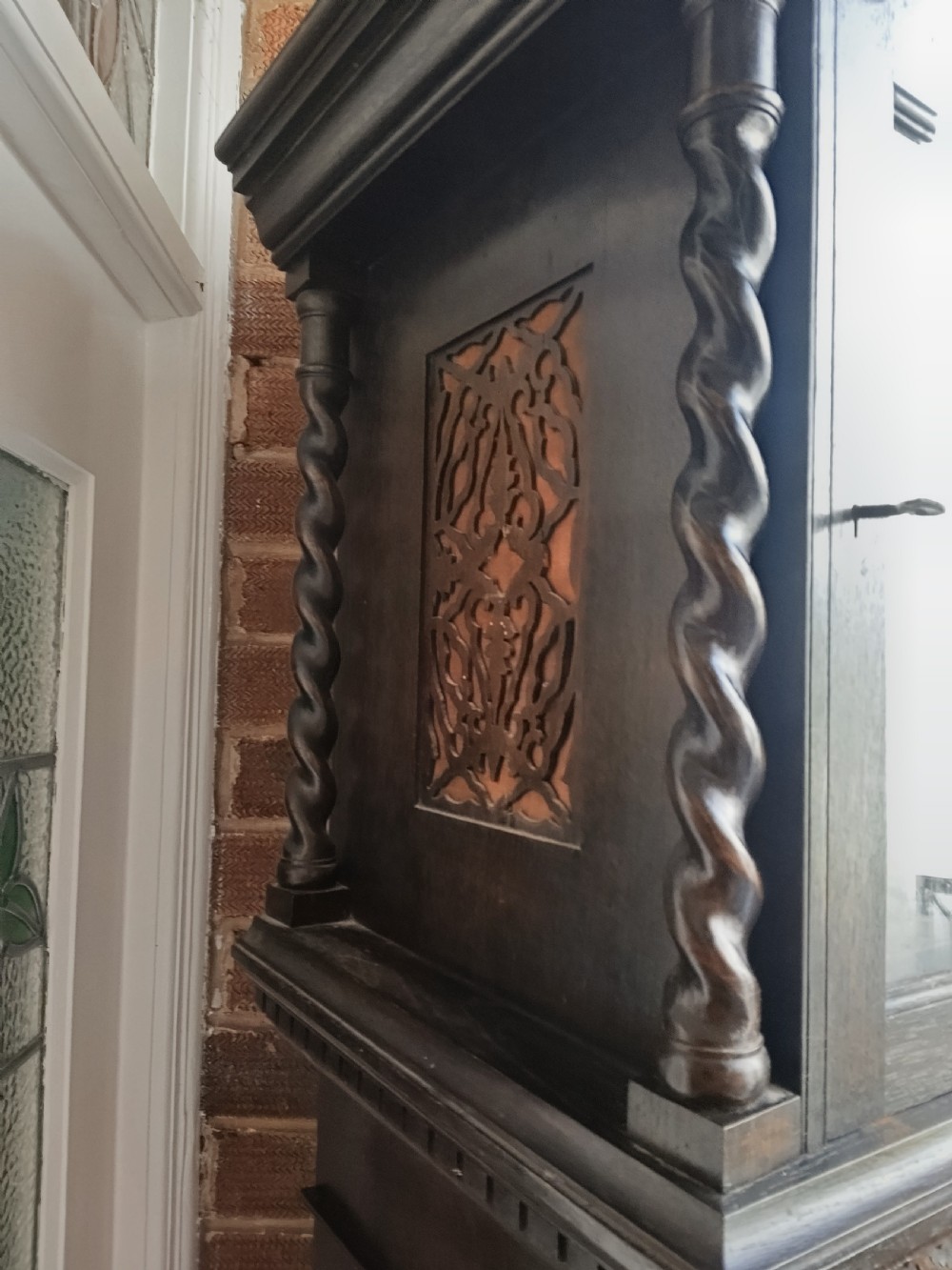 A tall dark oak longcase clock featuring an arched hood, barley twist columns, carved wood panels, and an ornate brass dial. View VII