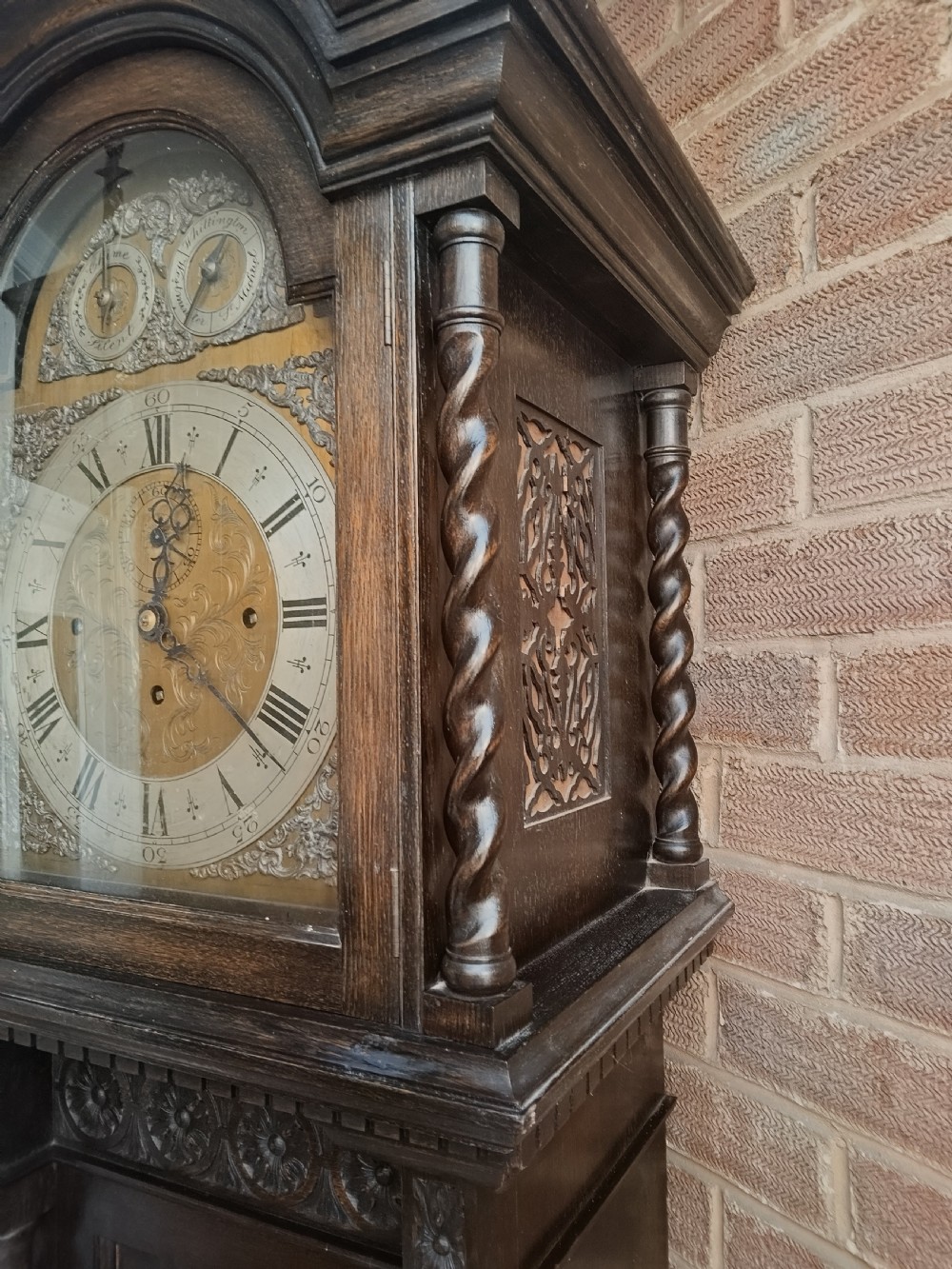 A tall dark oak longcase clock featuring an arched hood, barley twist columns, carved wood panels, and an ornate brass dial. View VIII