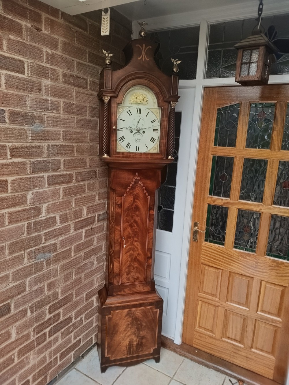 An antique flame mahogany longcase clock featuring a swan-neck pediment, brass finials, reeded columns, and a painted clock face with Roman numerals.
