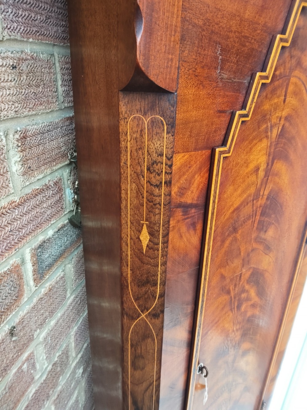 An antique flame mahogany longcase clock featuring a swan-neck pediment, brass finials, reeded columns, and a painted clock face with Roman numerals. View IV