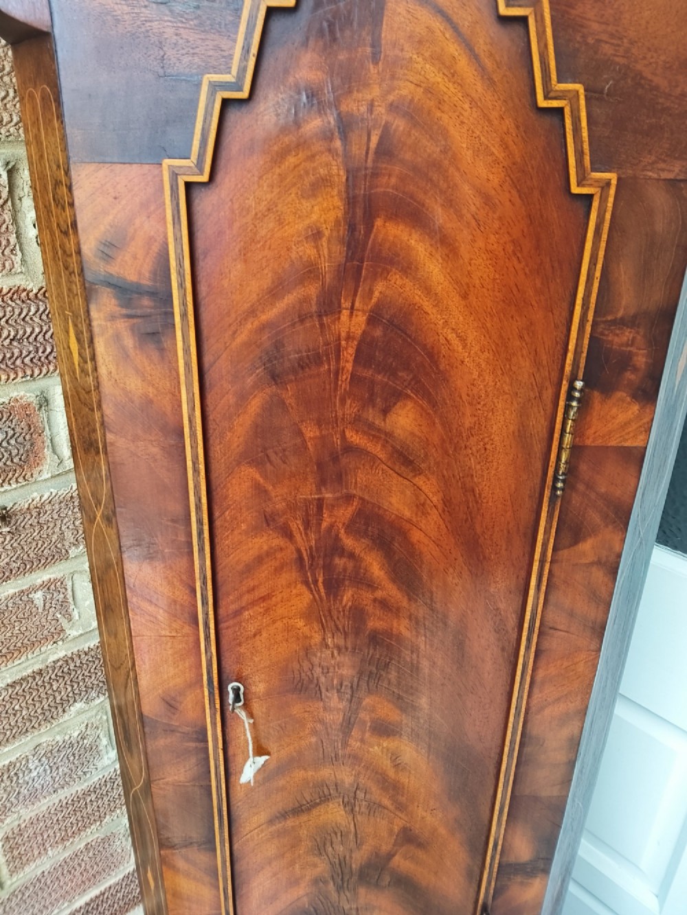 An antique flame mahogany longcase clock featuring a swan-neck pediment, brass finials, reeded columns, and a painted clock face with Roman numerals. View V