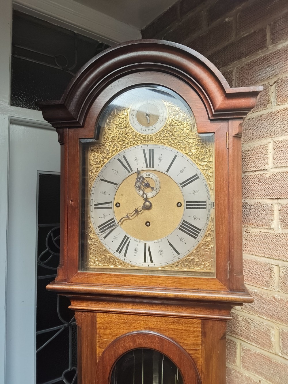 A tall wooden longcase clock featuring an arched hood, silvered brass dial, and a glazed door showcasing internal brass weights and pendulum. View II