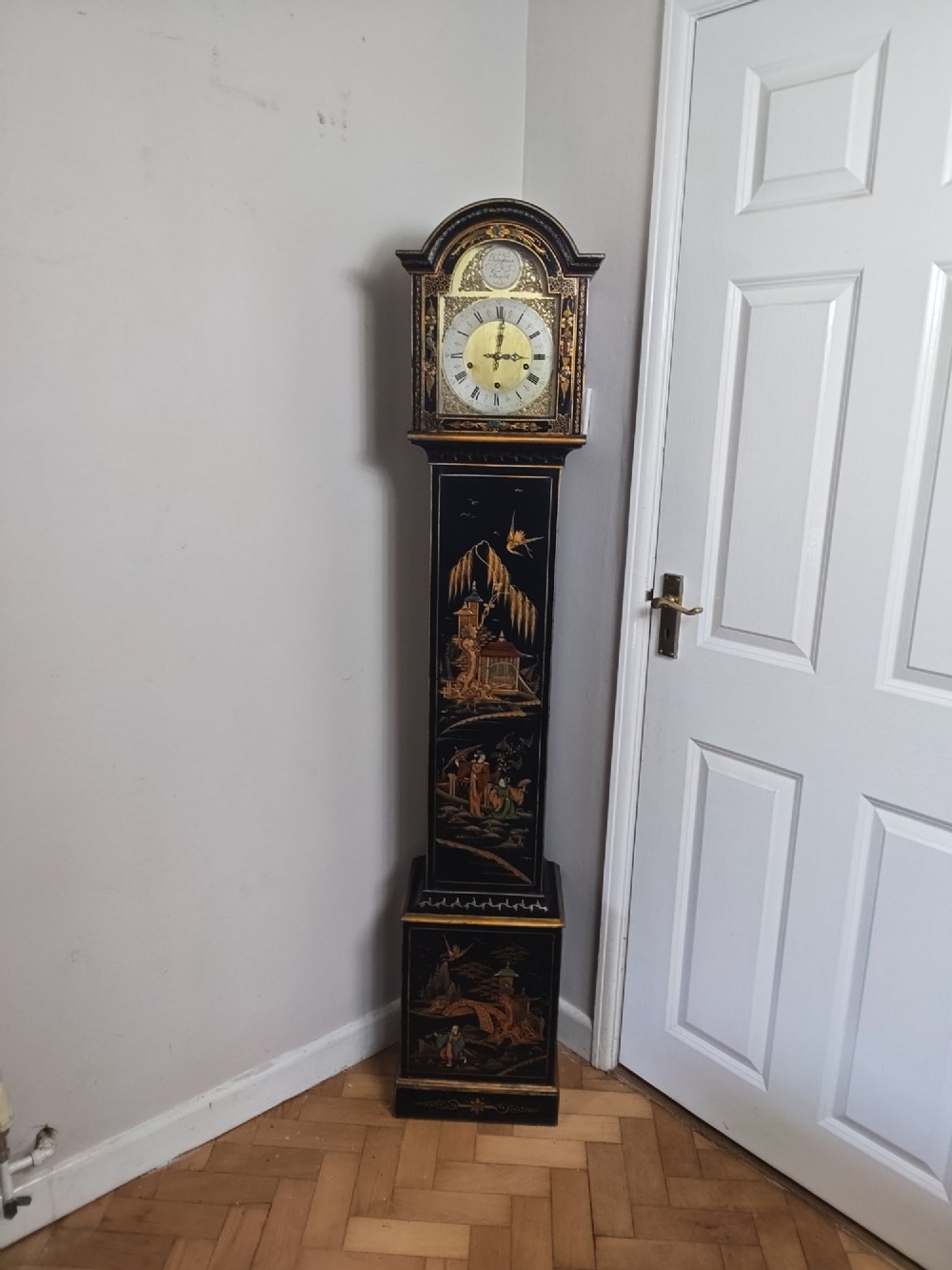 A slender black lacquer grandmother clock decorated with gilded chinoiserie landscape motifs and fitted with an arched brass dial.
