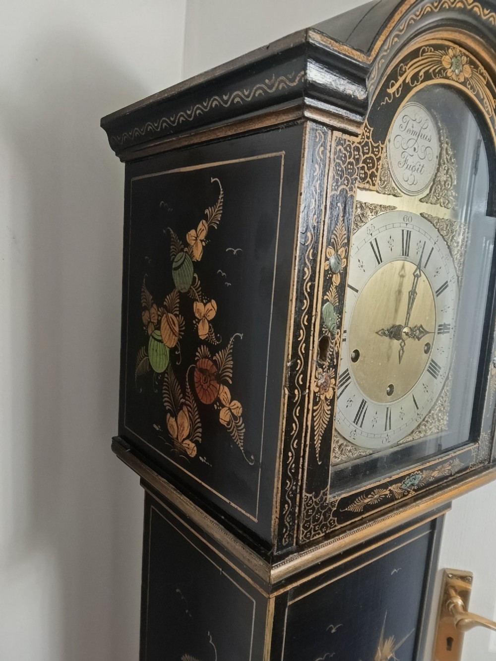 A slender black lacquer grandmother clock decorated with gilded chinoiserie landscape motifs and fitted with an arched brass dial. View V