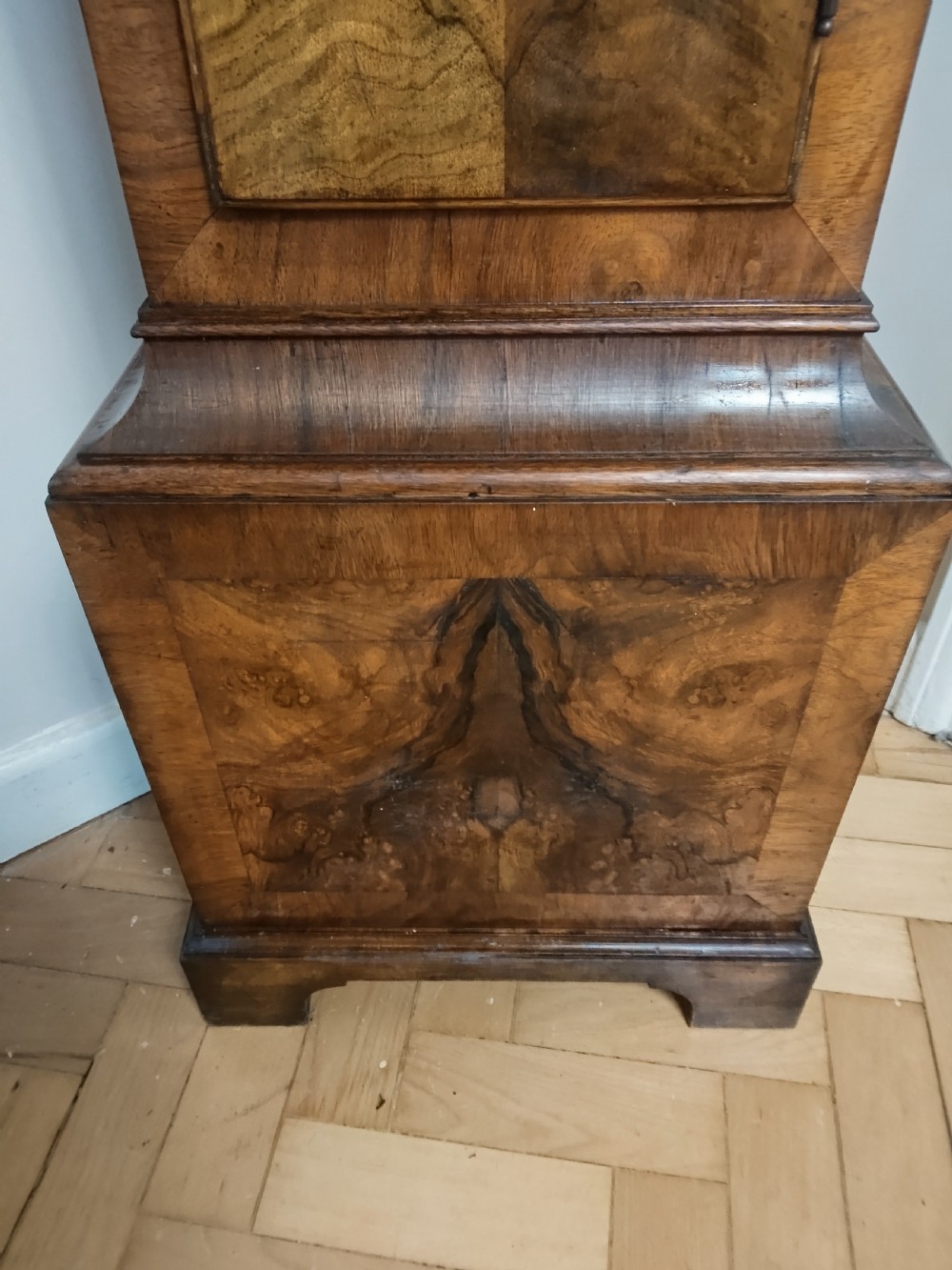 A slim burr walnut grandmother longcase clock featuring an arched hood, round silvered dial, and bookmatched timber grain. View IV