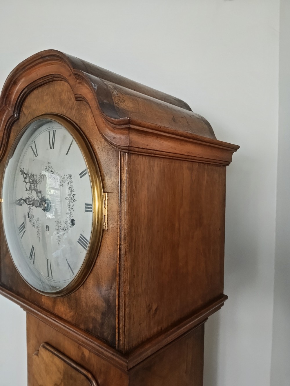 A slim burr walnut grandmother longcase clock featuring an arched hood, round silvered dial, and bookmatched timber grain. View V