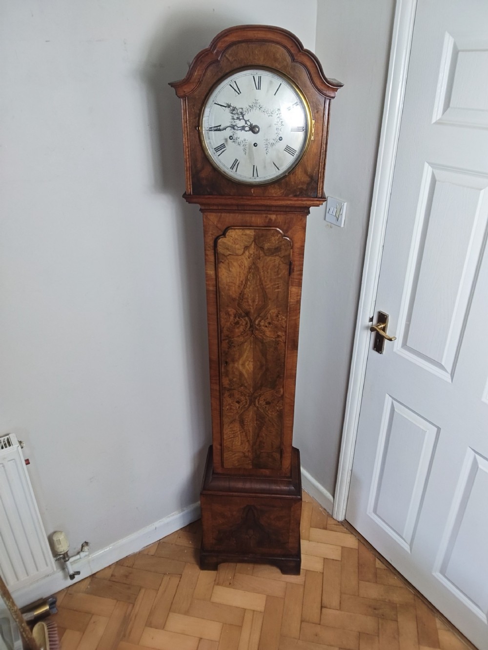 A slim burr walnut grandmother longcase clock featuring an arched hood, round silvered dial, and bookmatched timber grain.