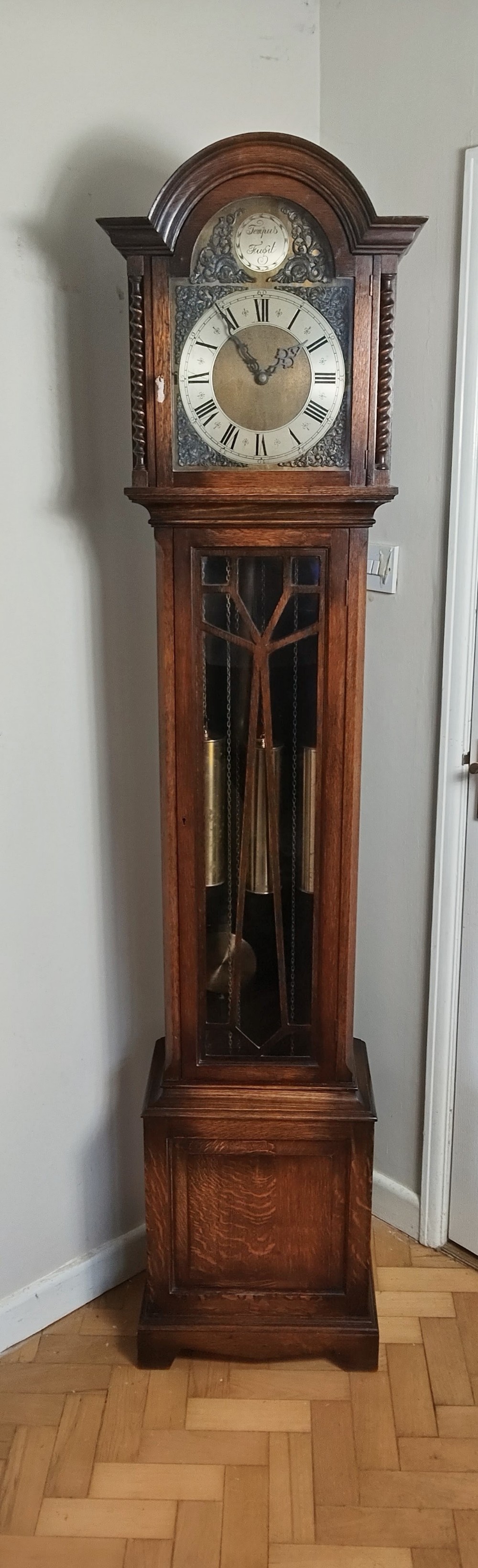A tall dark oak longcase clock featuring an arched bonnet, barley twist side columns, a brass dial with Roman numerals, and a glazed trunk door displaying three brass weights and pendulum.