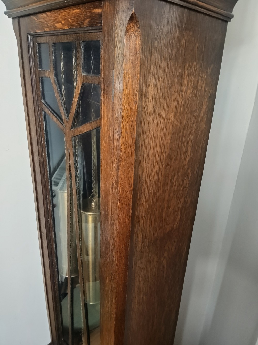 A tall dark oak longcase clock featuring an arched bonnet, barley twist side columns, a brass dial with Roman numerals, and a glazed trunk door displaying three brass weights and pendulum. View V